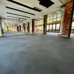 Wnętrze remontowanego biura z szarą wylewką samopoziomującą, odsłoniętymi ceglanymi filarami i niedokończonym sufitem.