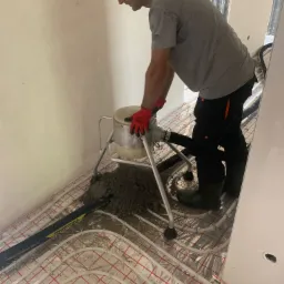 Pracownik w czerwonych rękawicach obsługuje maszynę do wylewania posadzki na ogrzewanie podłogowe w pomieszczeniu w stanie surowym.