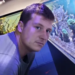 Mężczyzna pozujący przed akwarium z koralowcami, widoczny fragment twarzy i górnej części ciała, akwarium z oświetleniem LED w tle.