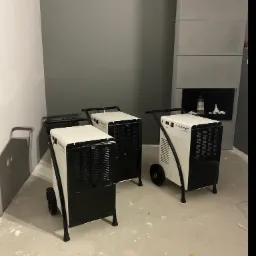 Trzy przemysłowe osuszacze powietrza marki Trotec ustawione w szarym pomieszczeniu z betonową podłogą i kominkiem w tle.