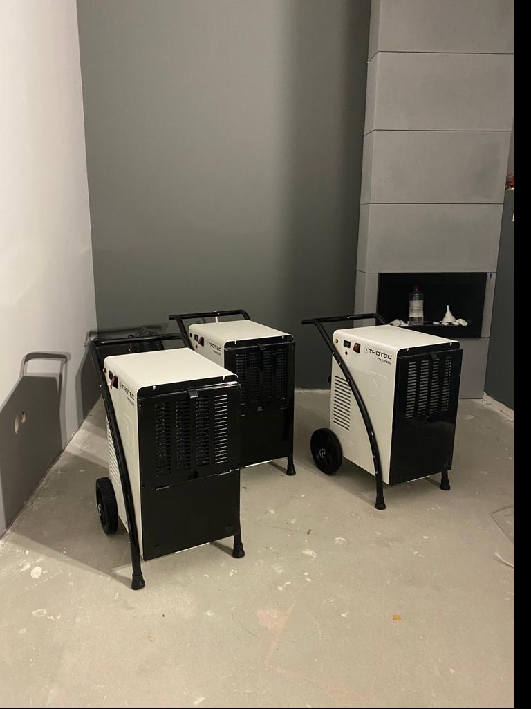 Trzy przemysłowe osuszacze powietrza marki Trotec ustawione w szarym pomieszczeniu z betonową podłogą i kominkiem w tle.
