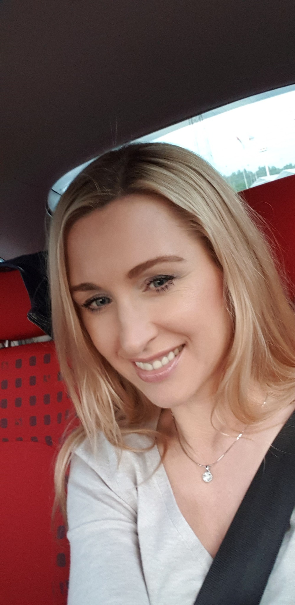 Uśmiechnięta blond kobieta z naszyjnikiem, siedząca w samochodzie z czerwoną tapicerką, patrzy w dół. Widoczne okno i pas bezpieczeństwa.