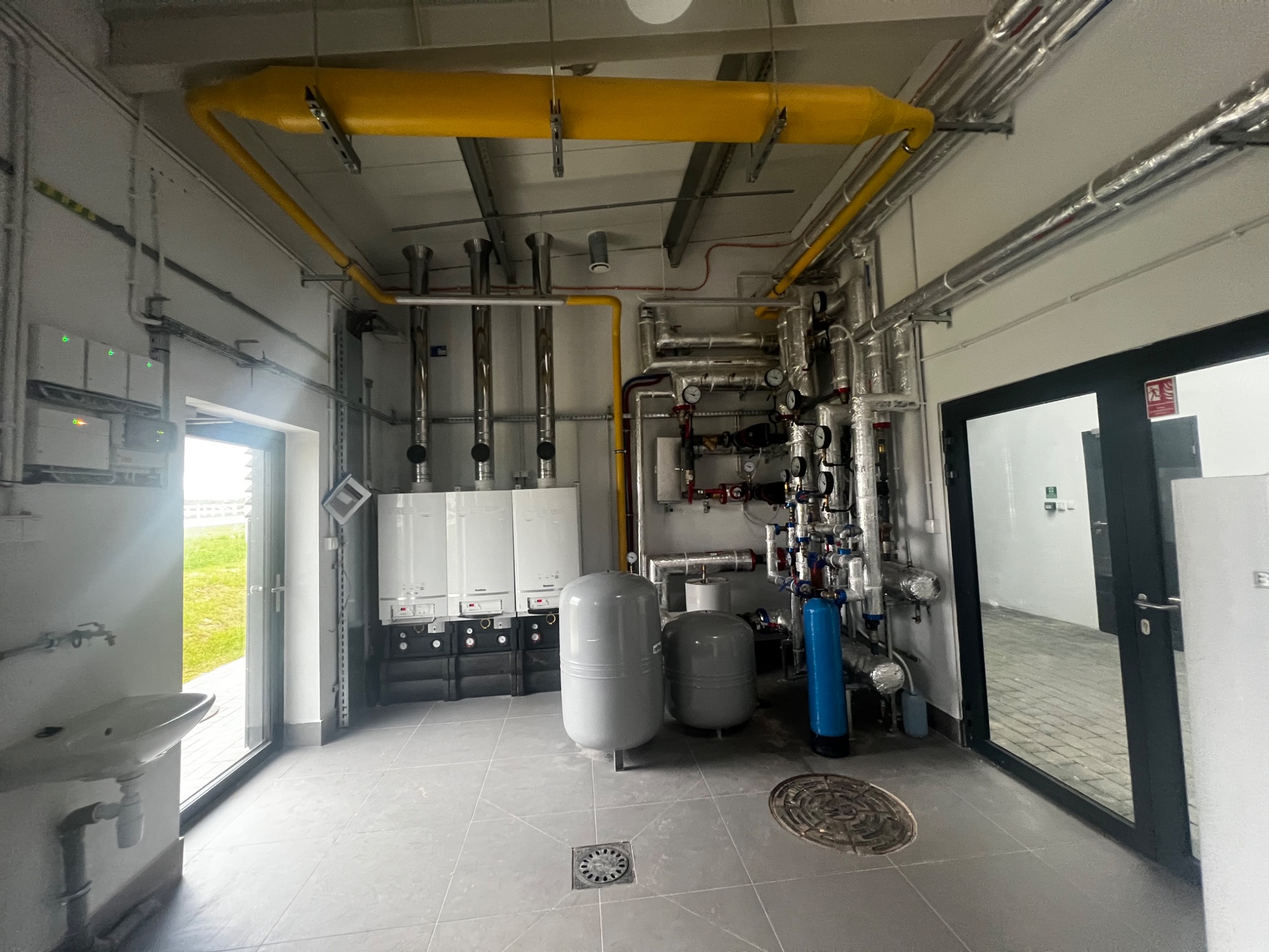 Kompletna instalacja kotłowni z dwoma kotłami grzewczymi, zbiornikami wyrównawczymi, filtrem wody i rozbudowaną armaturą, widoczna umywalka w pomieszczeniu z szarymi płytkami podłogowymi.