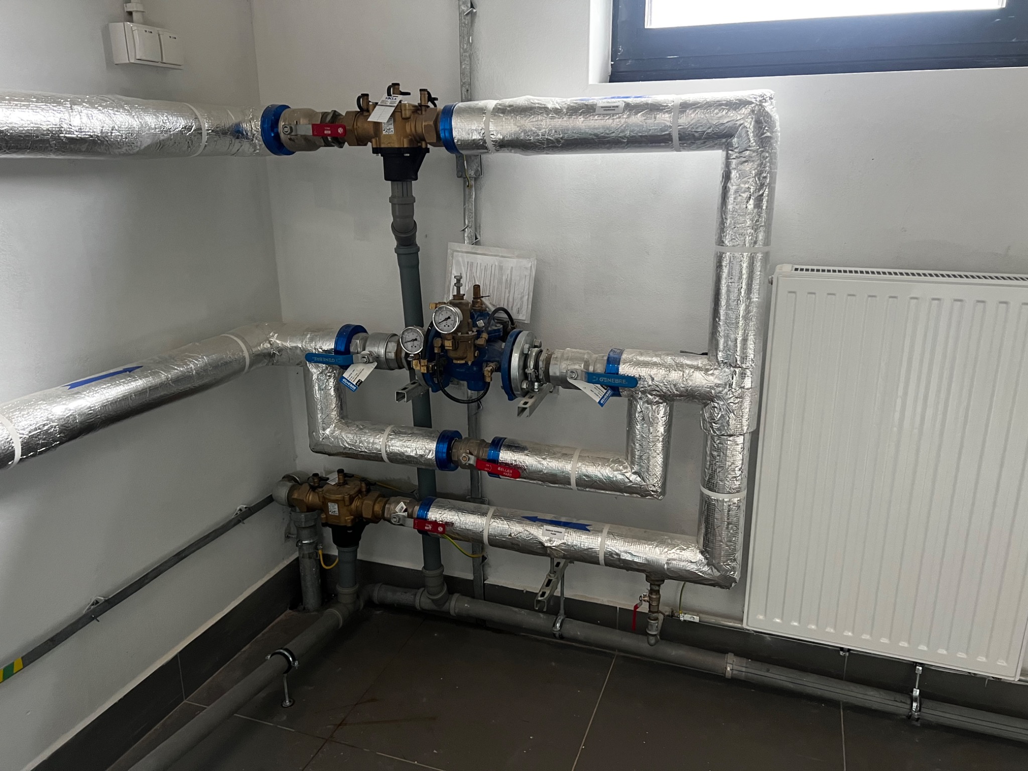 Izolowane rury z zaworami i manometrami w pomieszczeniu technicznym, widoczny grzejnik i fragment okna.