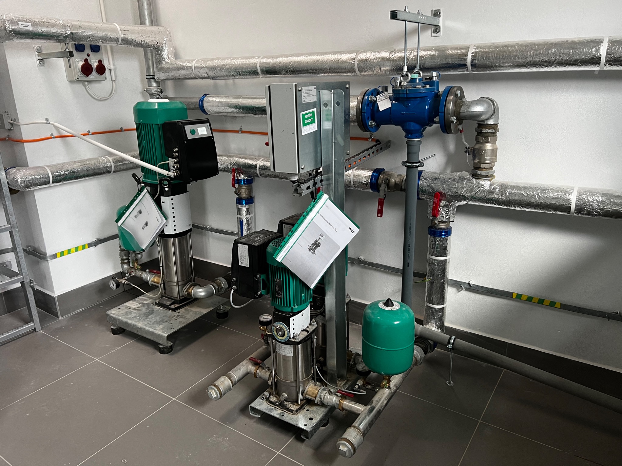 Dwie identyczne, zainstalowane pompy obok siebie w pomieszczeniu technicznym z szarymi płytkami na podłodze i izolowanymi rurami na ścianach, z dokumentacją techniczną zawieszoną na jednej z nich.