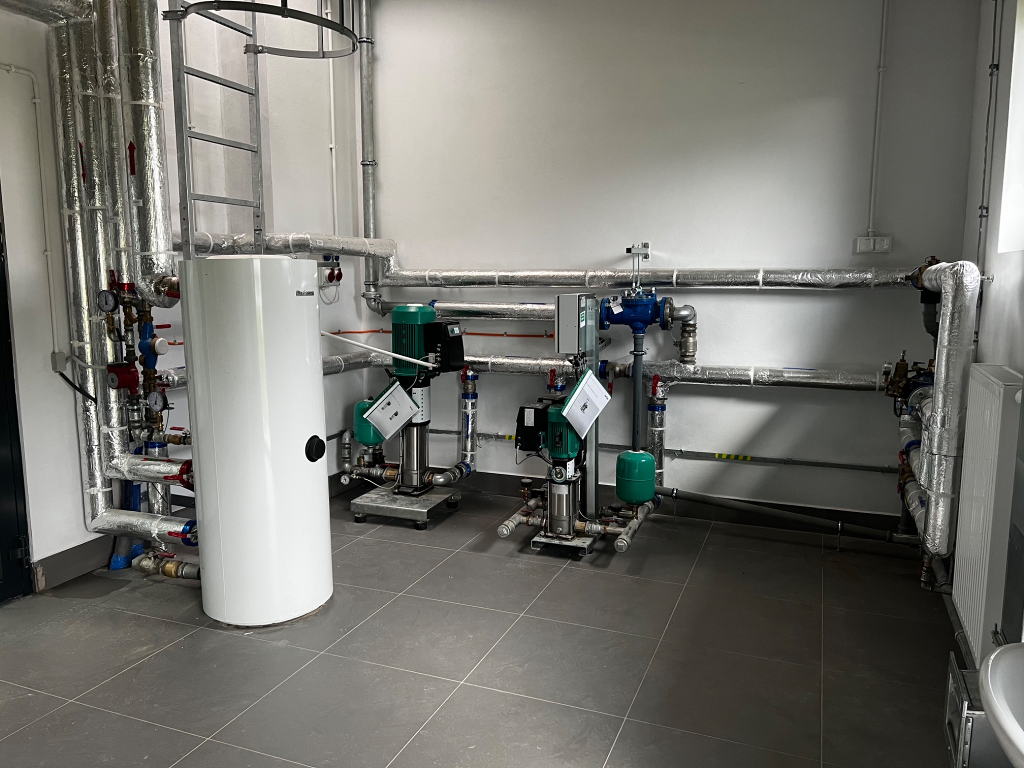 Instalacja z dwoma pompami obiegowymi, zbiornikiem akumulacyjnym i rurami w izolacji w pomieszczeniu technicznym z szarą podłogą i drabinką przy ścianie.