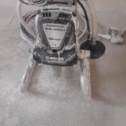 Używany agregat malarski marki Mar-Pol, model M80636, z widocznymi śladami farby na obudowie, stojący na podłodze pokrytej pyłem, z przewodem i pistoletem natryskowym w tle.