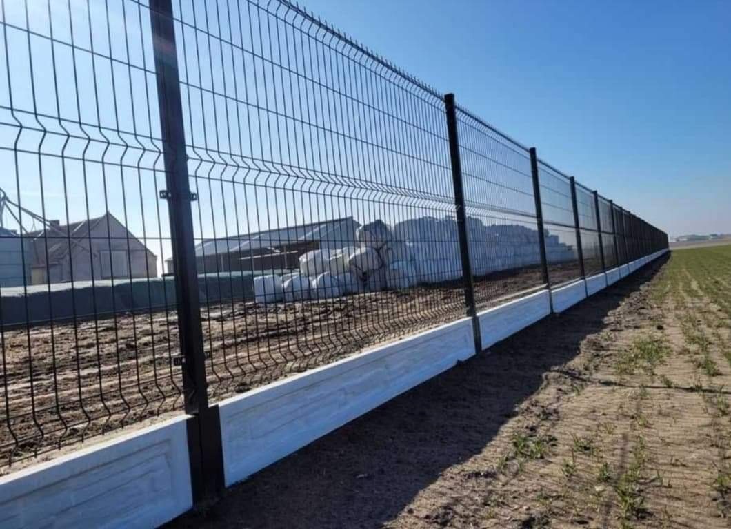 Długi, czarny panelowy płot z betonową podmurówką, rozciągający się wzdłuż pola w słoneczny dzień, z budynkami gospodarczymi i stertą worków w tle.