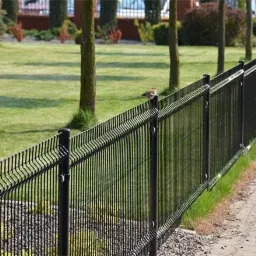 Długa, czarna, metalowa siatka ogrodzeniowa z drobnymi oczkami, ciągnąca się wzdłuż ścieżki, z małym brązowym ptakiem siedzącym na jej szczycie, trawnik i drzewa w tle.