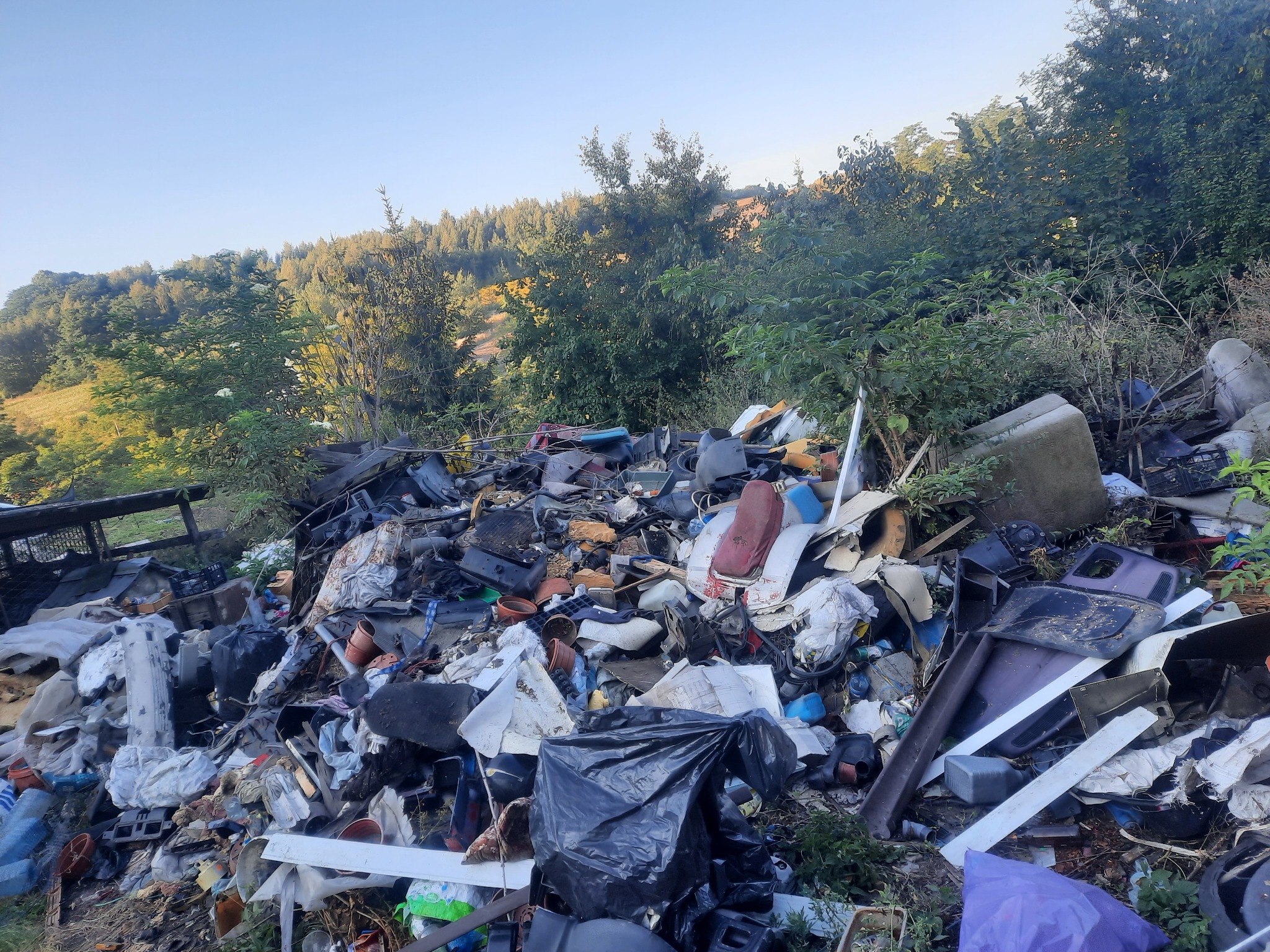 Nielegalne wysypisko śmieci w Krakowie: sterta różnorodnych odpadów, w tym elementy plastikowe, części samochodowe i worki na śmieci, na tle zieleni drzew i krzewów.