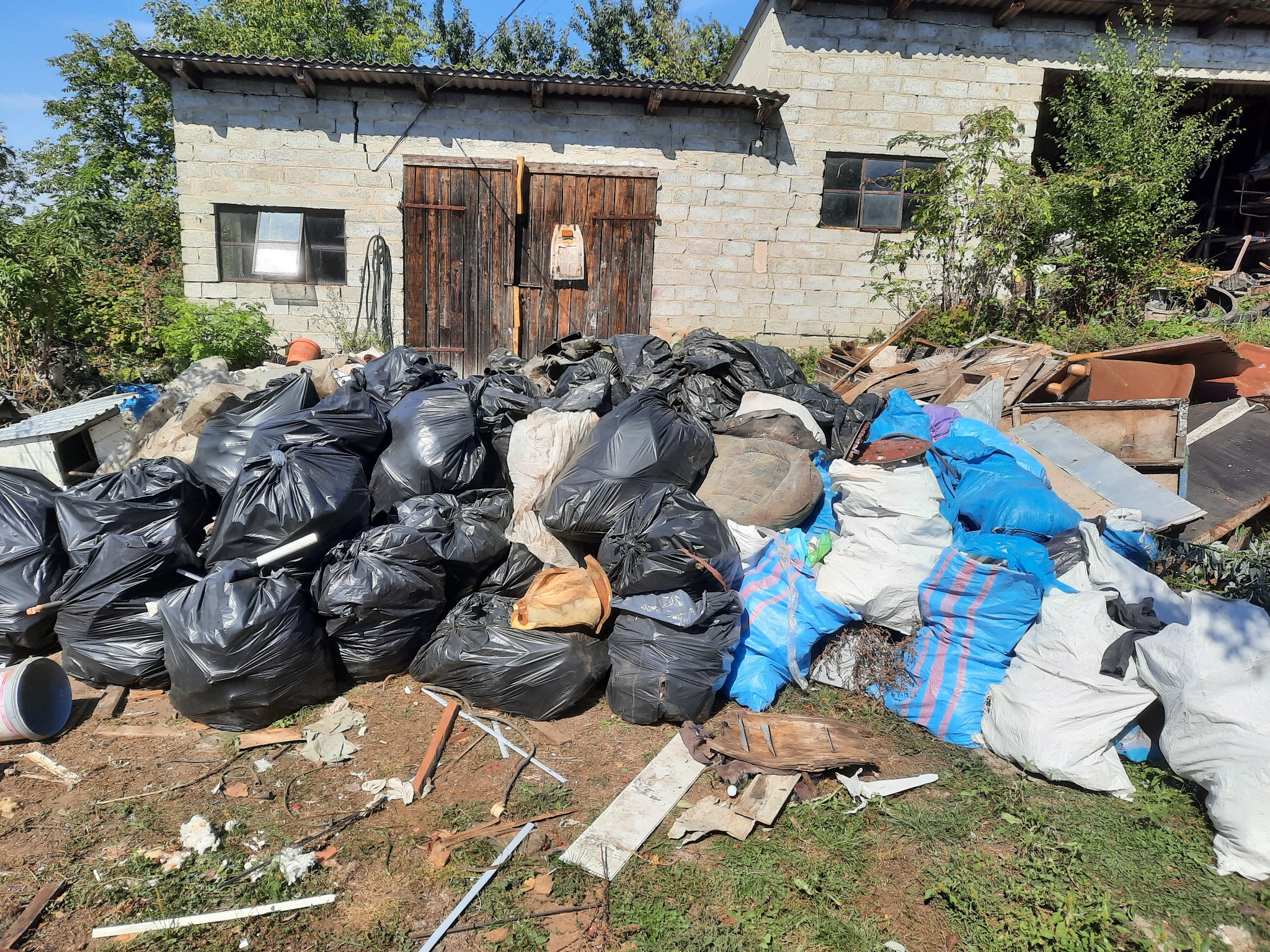 Sterta worków z gruzem, czarnych i niebieskich, zgromadzona przed murowanym budynkiem gospodarczym z drewnianymi wrotami i dwoma oknami.
