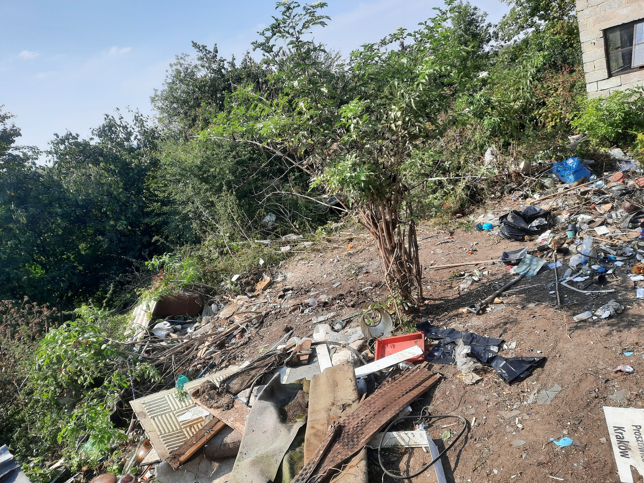 Dzikie wysypisko śmieci na zboczu z widocznymi gruzami, odpadami plastikowymi i workami na śmieci, porośnięte drzewami i krzewami, z fragmentem budynku w tle.