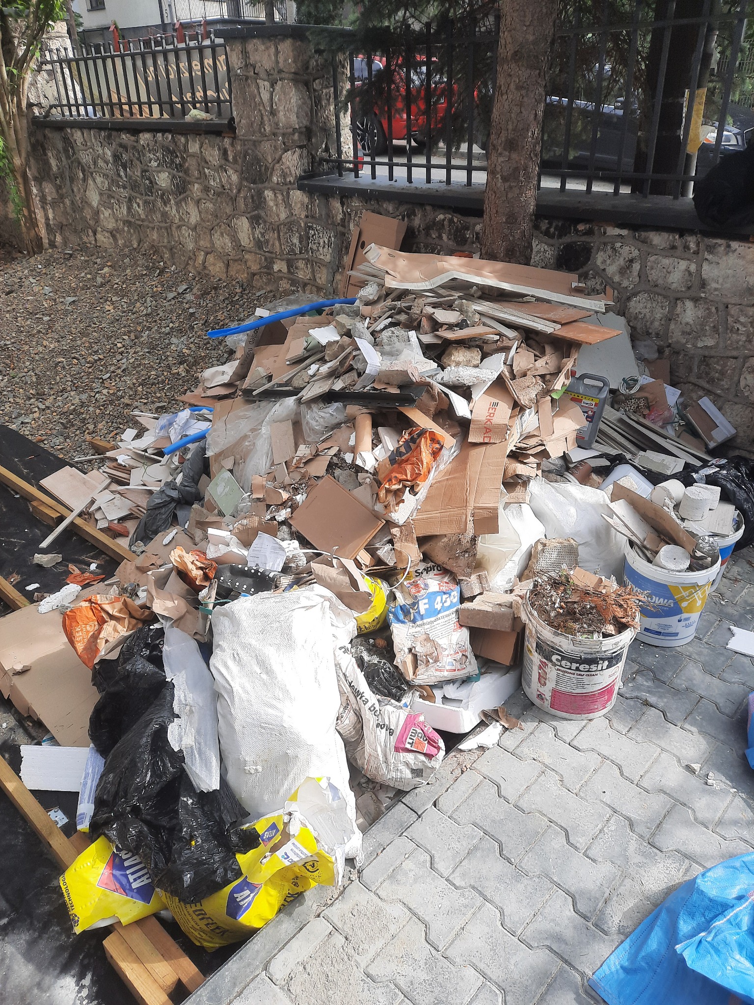 Stos gruzu i odpadów budowlanych na chodniku przy kamiennym murku z metalowym ogrodzeniem, widoczne fragmenty kartonów, worków, wiader po farbach i zaprawach, oraz niebieski fragment folii.