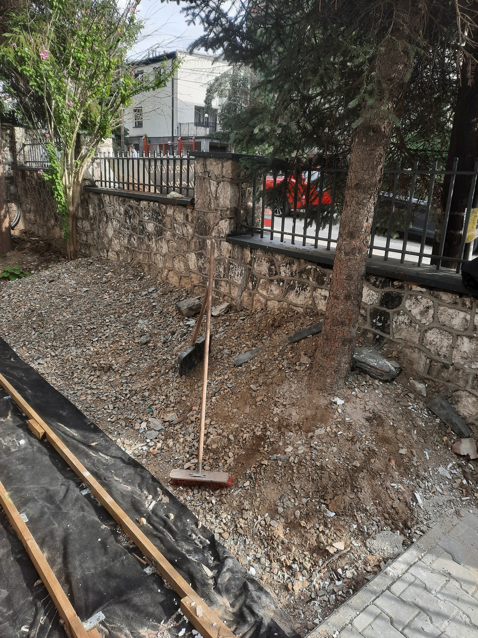 Przygotowanie terenu pod budowę: rozłożona czarna folia, drewniane belki, rozsypany gruz, grabie oparte o ścianę z kamienia, drzewo iglaste obok.