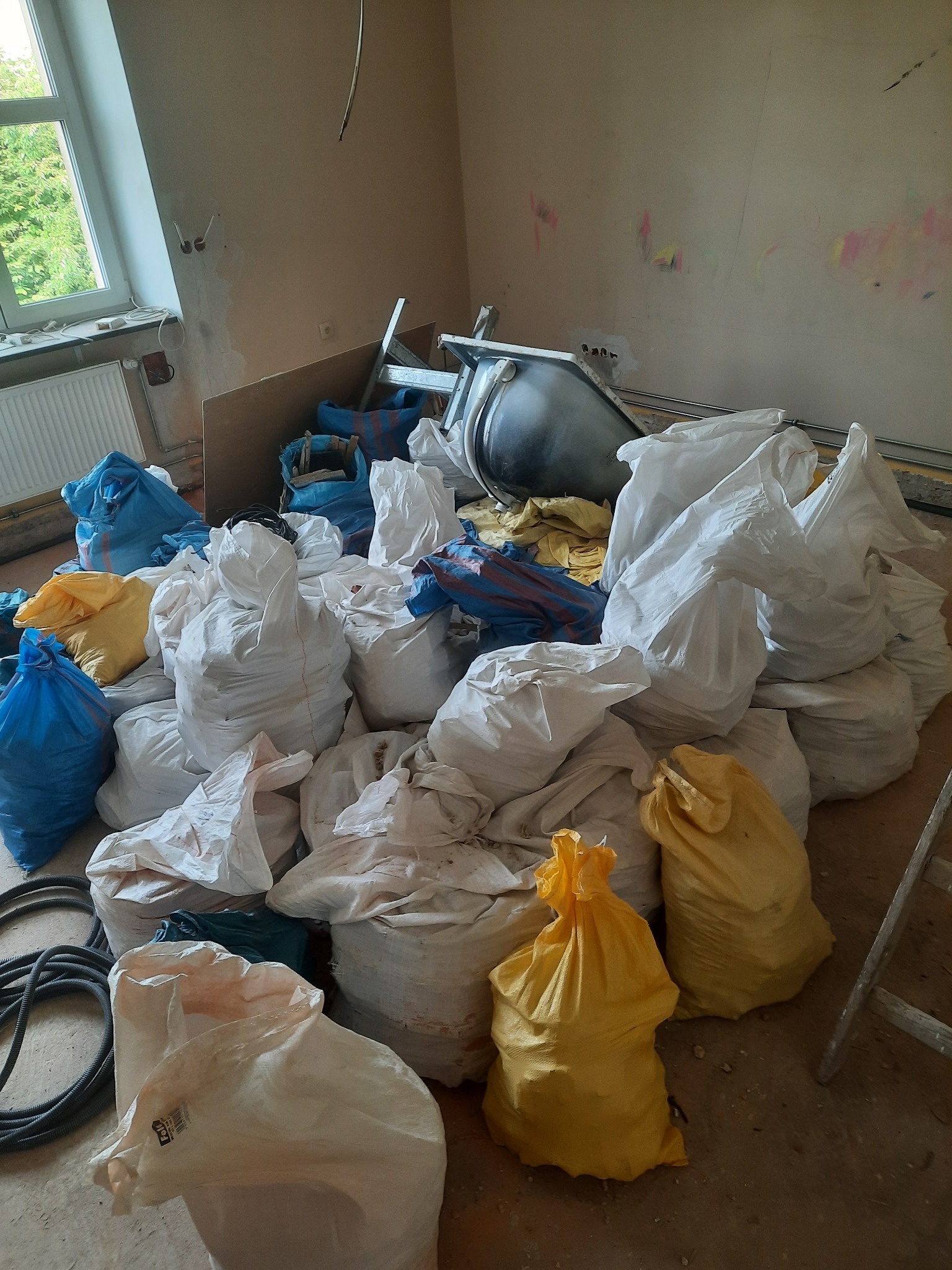 Stos worków z gruzem w pomieszczeniu w trakcie remontu, widoczna wanna, drabina i przewody elektryczne, ściany z śladami po instalacjach.