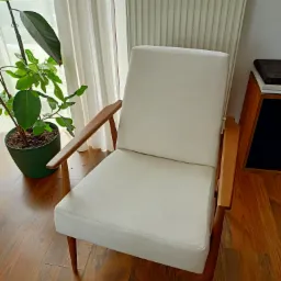 Biały fotel z drewnianymi podłokietnikami i nogami, stojący na parkiecie obok zielonej rośliny w doniczce i grzejnika