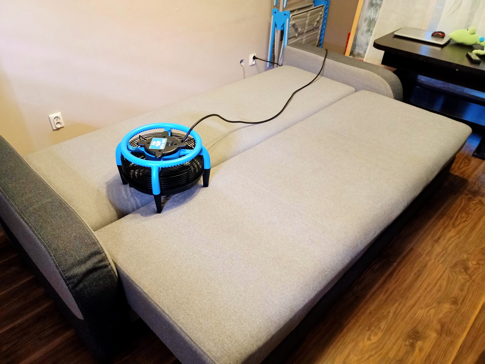 Rozłożona szara kanapa z podłączonym do gniazdka wentylatorem do osuszania tapicerki, widoczne ślady po czyszczeniu na podłokietniku.