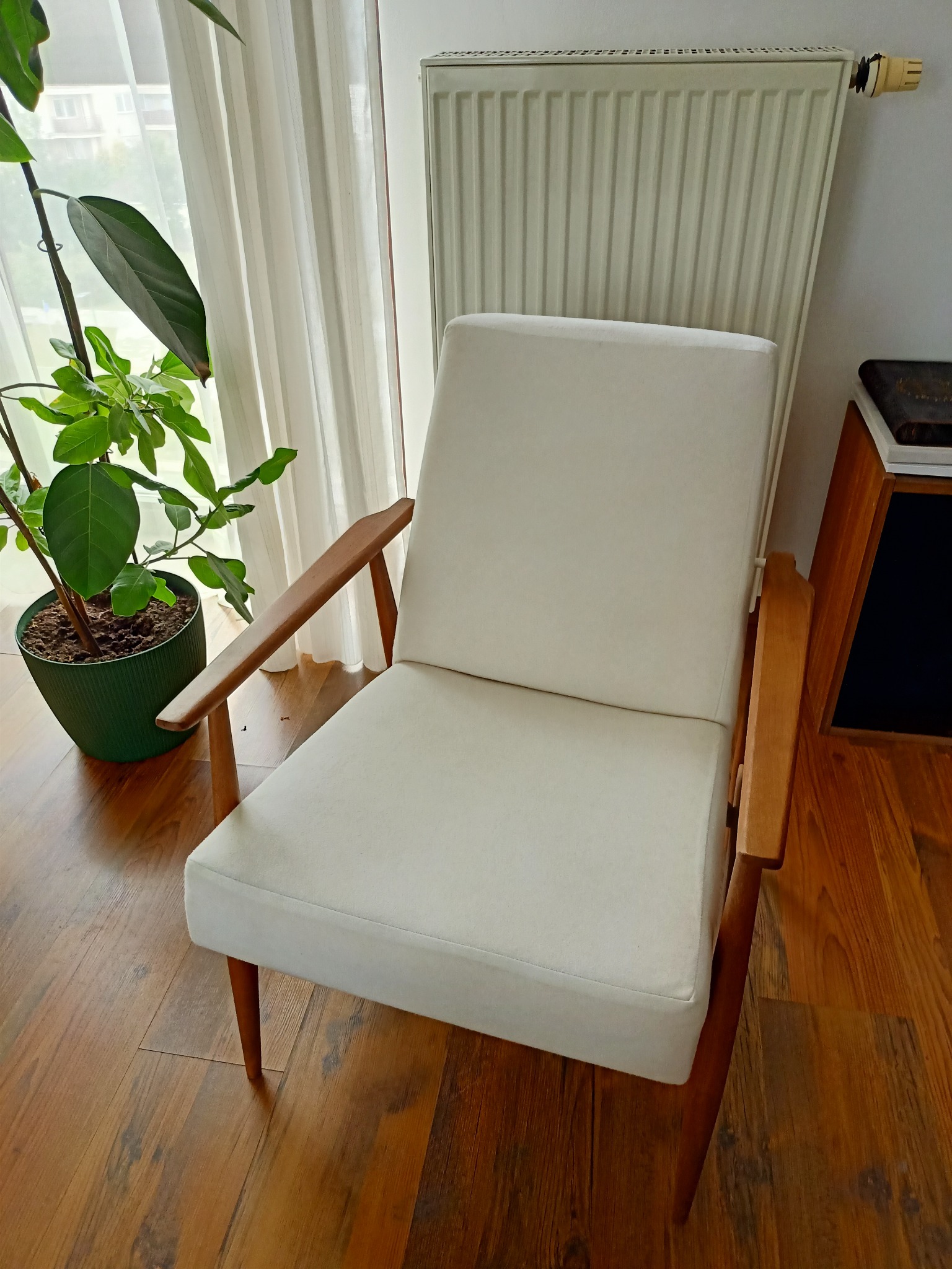 Biały fotel z drewnianymi podłokietnikami i nogami, stojący na parkiecie obok zielonej rośliny w doniczce i grzejnika