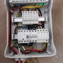 Otwarta, biała skrzynka elektryczna z widocznymi bezpiecznikami, przewodami w różnych kolorach (czerwony, żółty, zielony, czarny, niebieski) oraz elementami montażowymi na szynach DIN.