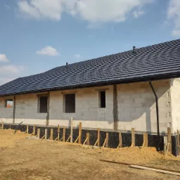 Dom w trakcie budowy z czarnym dachem z blachy na tle błękitnego nieba. Widoczne rynny i kominy. Fundamenty zabezpieczone folią i drewnianymi podporami.