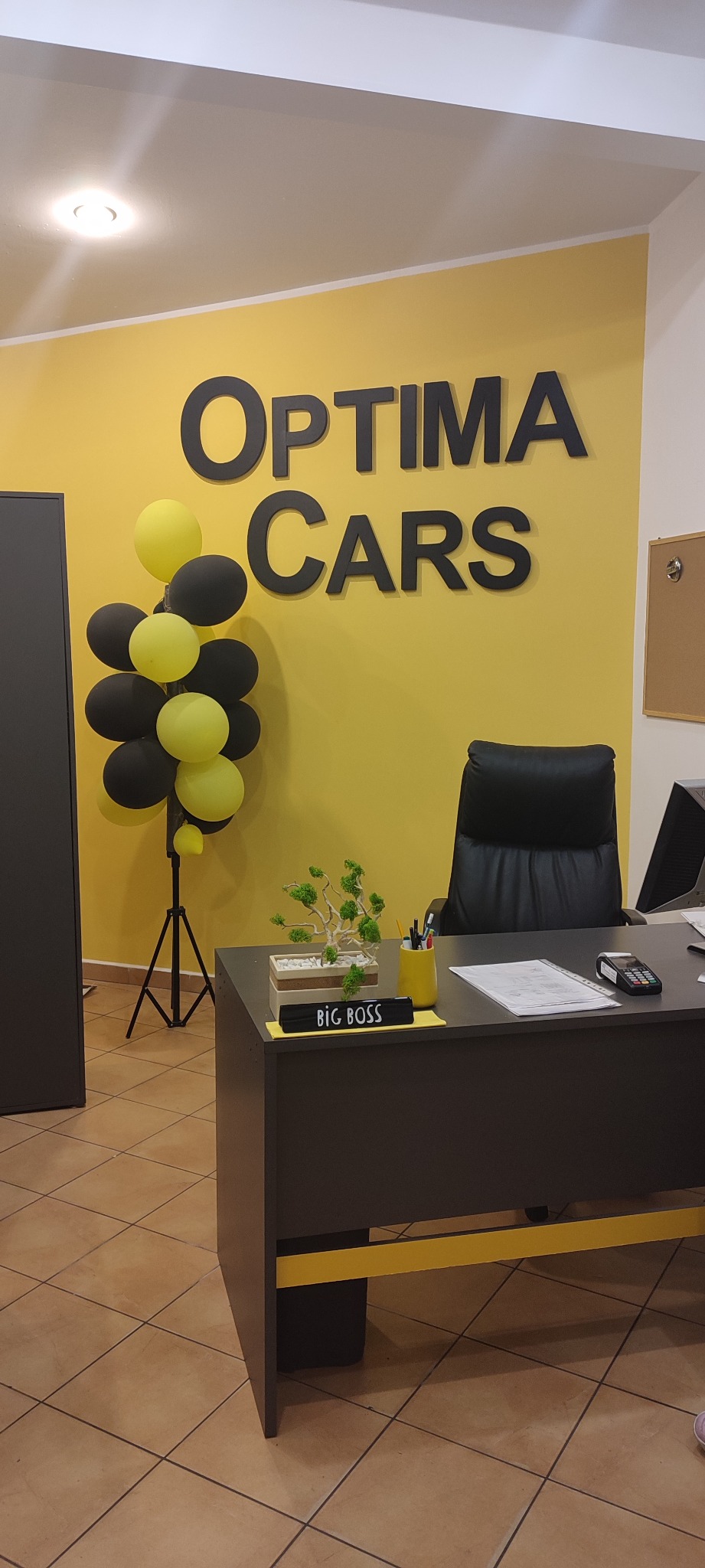 Biuro firmy Optima Cars z żółtą ścianą, czarnymi literami logo, balonami w kolorach żółtym i czarnym, biurkiem z napisem 'Big Boss' oraz rośliną w doniczce.