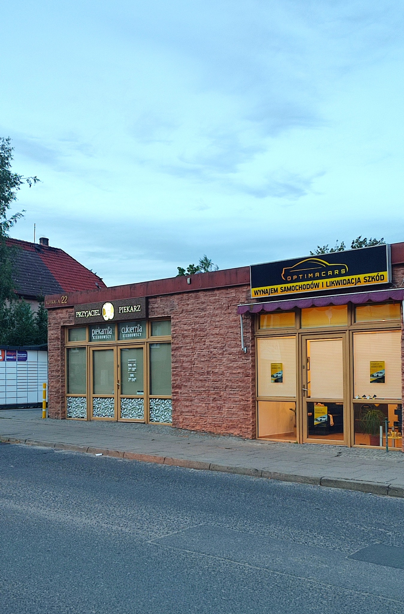 Budynek z dwoma lokalami: piekarnia/cukiernia oraz wypożyczalnia samochodów z napisem 'Wynajem samochodów i likwidacja szkód' na szyldzie.
