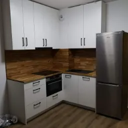 Nowoczesna kuchnia z białymi szafkami, drewnianym blatem i brązowym panelem nad blatem, srebrna lodówka, widoczne opakowania po montażu na podłodze.