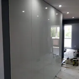 Błyszcząca, biała szafa wnękowa z oświetleniem punktowym w suficie, widoczna podczas montażu w Oławie, z narzędziami i materiałami zabezpieczającymi podłogę.