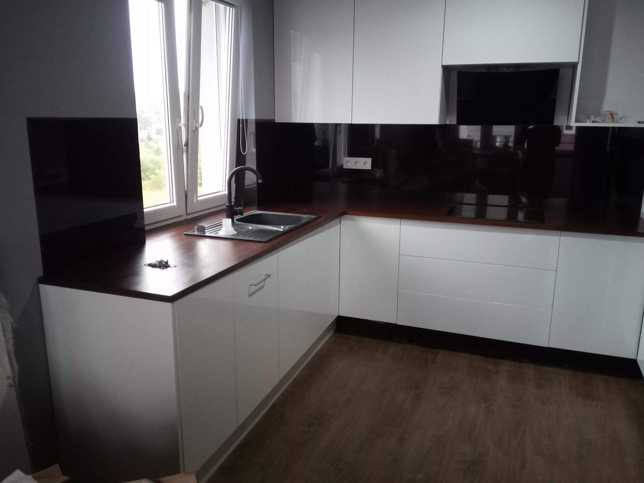 Nowoczesna kuchnia z białymi, lakierowanymi frontami, ciemnym blatem i czarnym panelem szklanym nad blatem, widok na okno z białymi ramami i zasłoną.