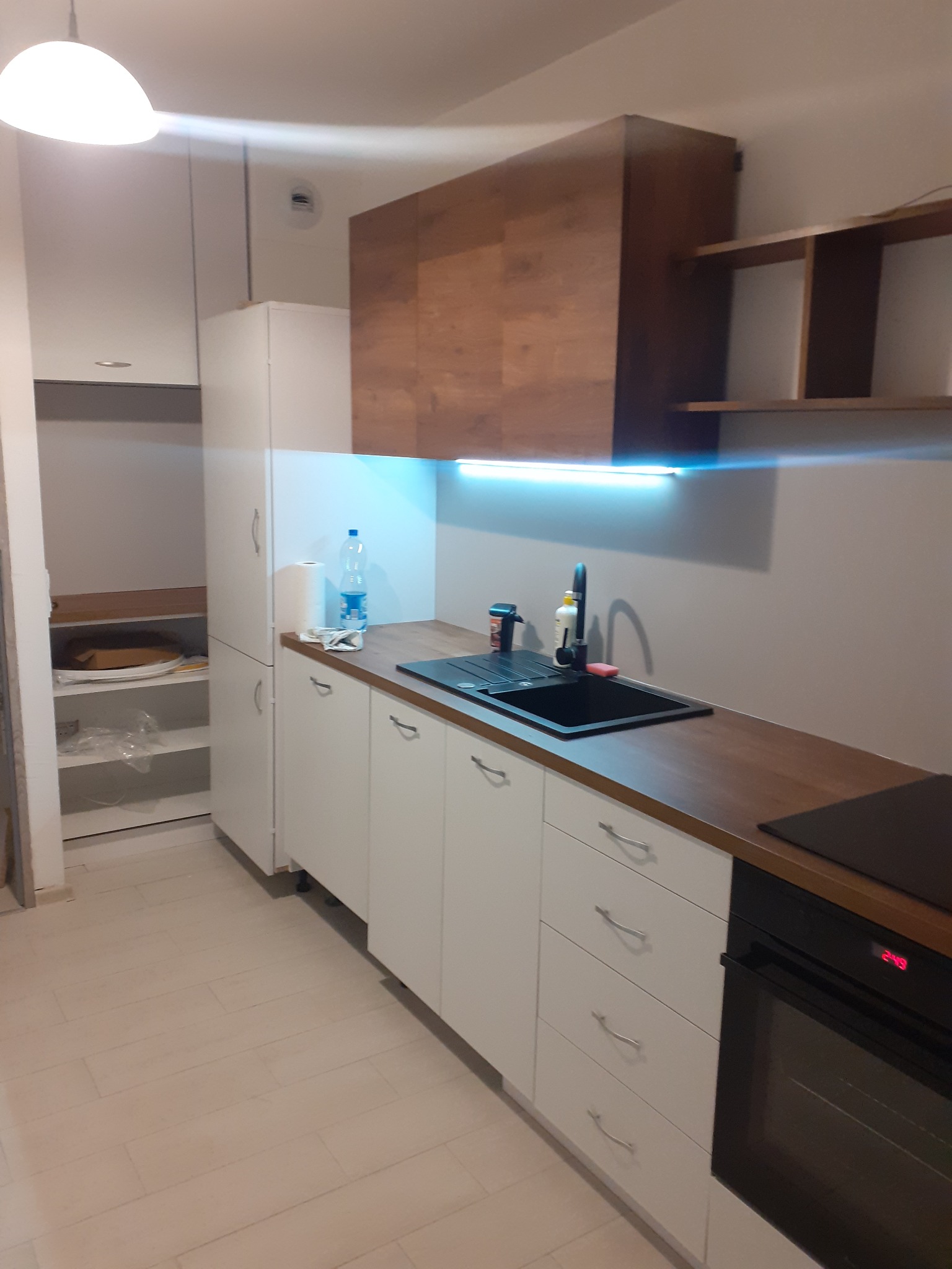 Nowoczesna kuchnia z białymi szafkami z szufladami i drewnianym blatem, czarny zlewozmywak z baterią, wisząca szafka z drewnianym frontem oraz oświetlenie LED podszafkowe.