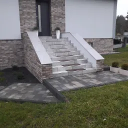 Wejście do domu z kostki brukowej i schodami wykończonymi płytkami, z donicami po bokach i na schodach, widok z trawnika.