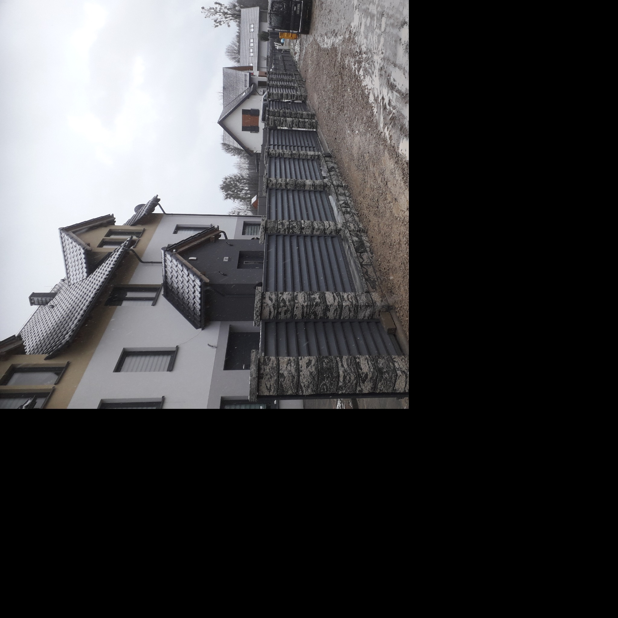 Szary, panelowy płot z kamiennymi słupkami oddzielający posesje, widok na domy w pochmurny dzień.