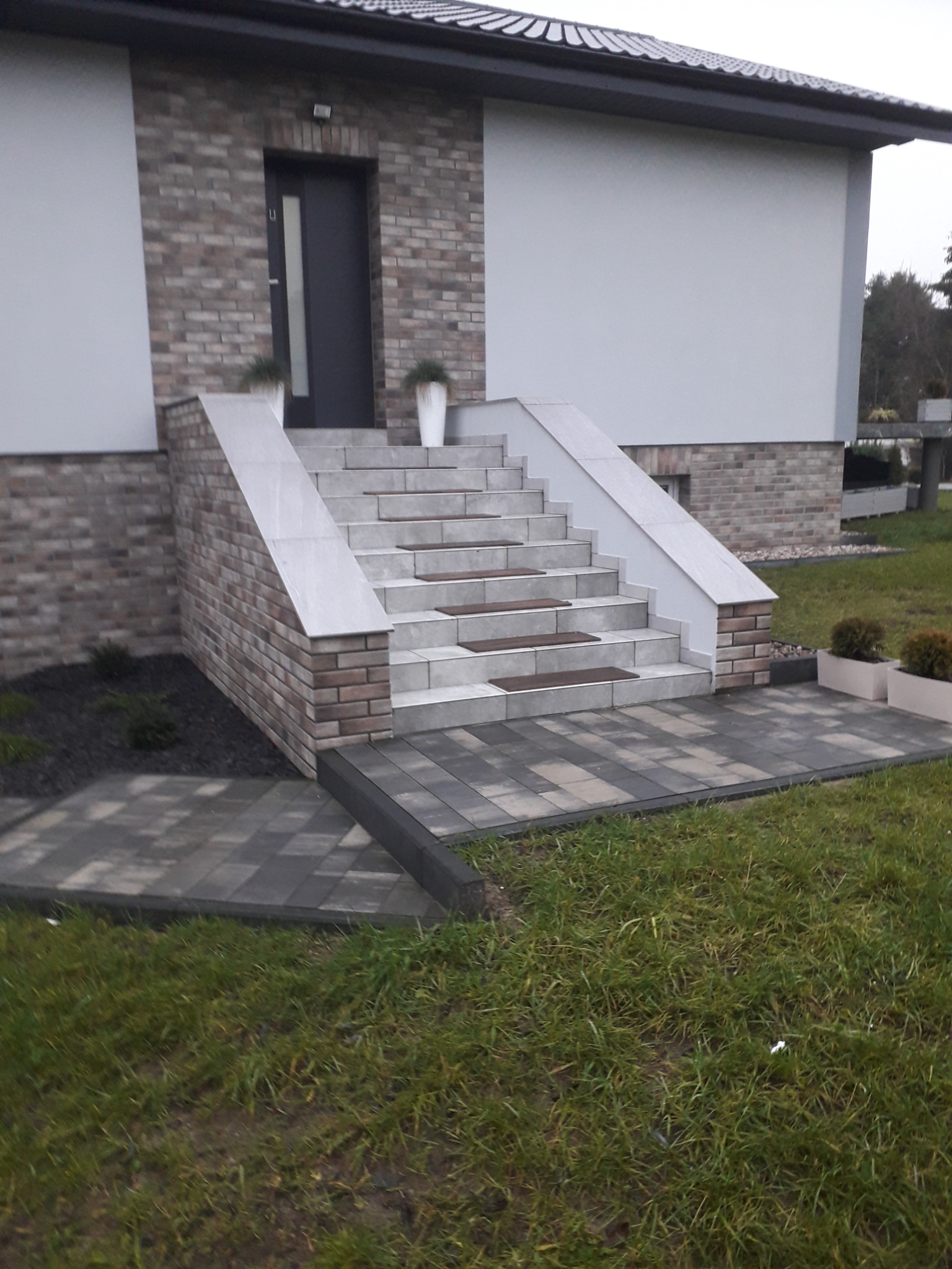 Wejście do domu z kostki brukowej i schodami wykończonymi płytkami, z donicami po bokach i na schodach, widok z trawnika.