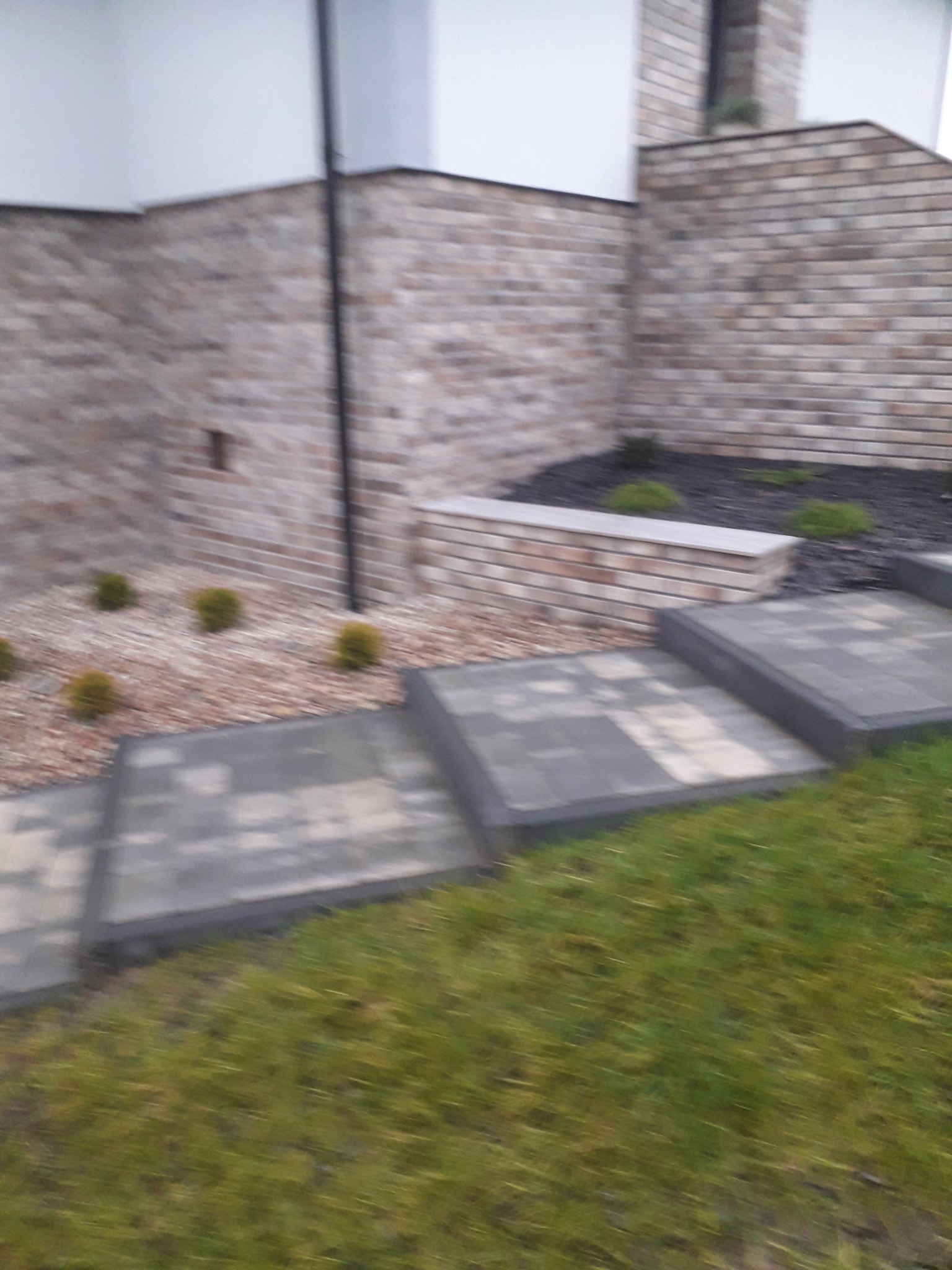 Schody z szarych płyt brukowych prowadzące do domu z ceglaną elewacją i rabatą z roślinami.