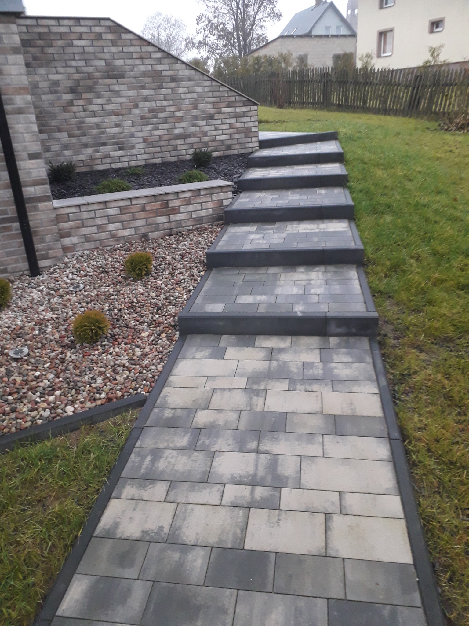 Schody z kostki brukowej prowadzące w górę, obok muru z cegły i kamiennego rabatu z roślinami.