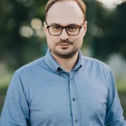 Portret mężczyzny w okularach z brązowymi oprawkami i błękitnej koszuli na tle rozmytej zieleni.