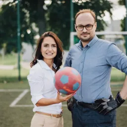 Uśmiechnięta kobieta trzyma piłkę do piłki nożnej na tle boiska, obok stoi mężczyzna w rękawicach bramkarskich, oboje w strojach biznesowych.