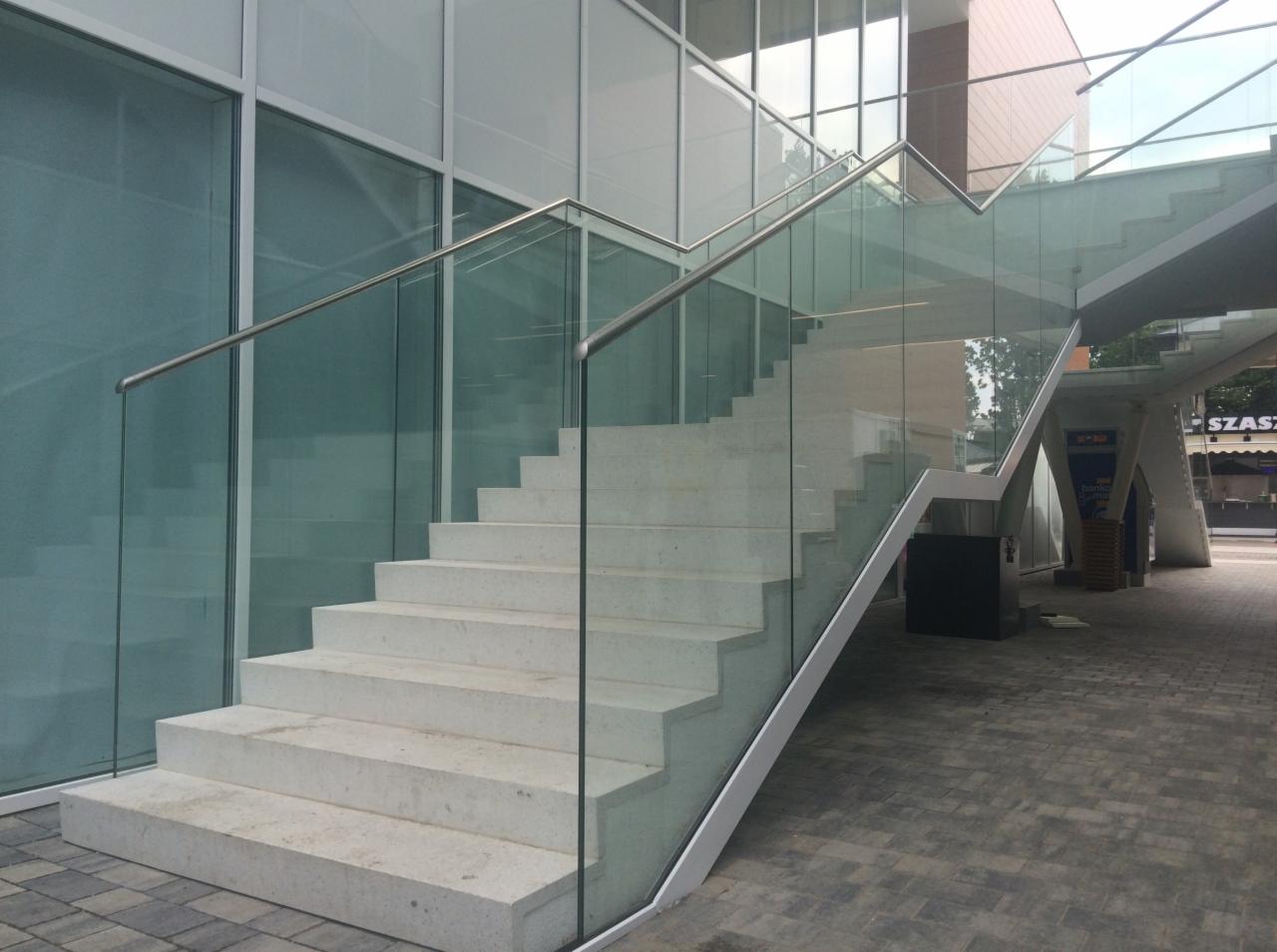 Nowoczesne schody zewnętrzne z białego kamienia z balustradą ze szkła i stali nierdzewnej, widoczne na tle przeszklonej fasady budynku.