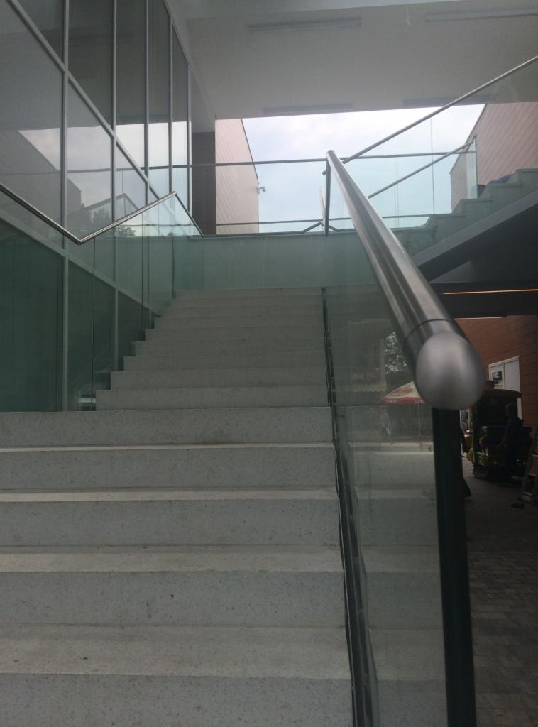 Schody z szarych płyt z przeszklonymi panelami i metalową poręczą, widok z dołu. Nowoczesna architektura z dużymi oknami w tle.