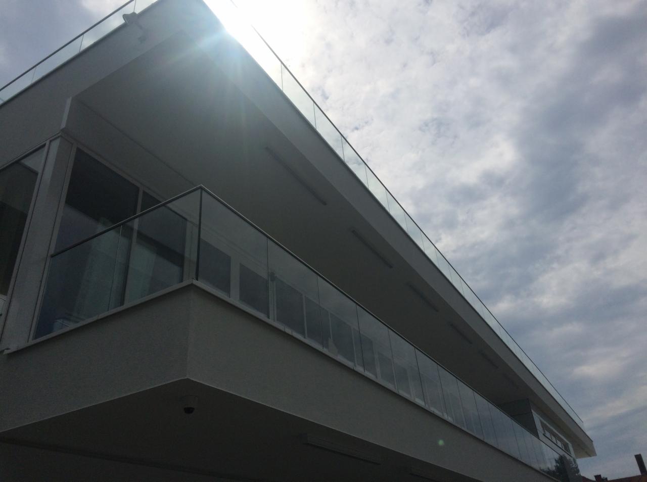 Nowoczesny budynek z balkonami zabezpieczonymi szklanymi panelami, widok z dołu w kierunku słonecznego nieba z chmurami.