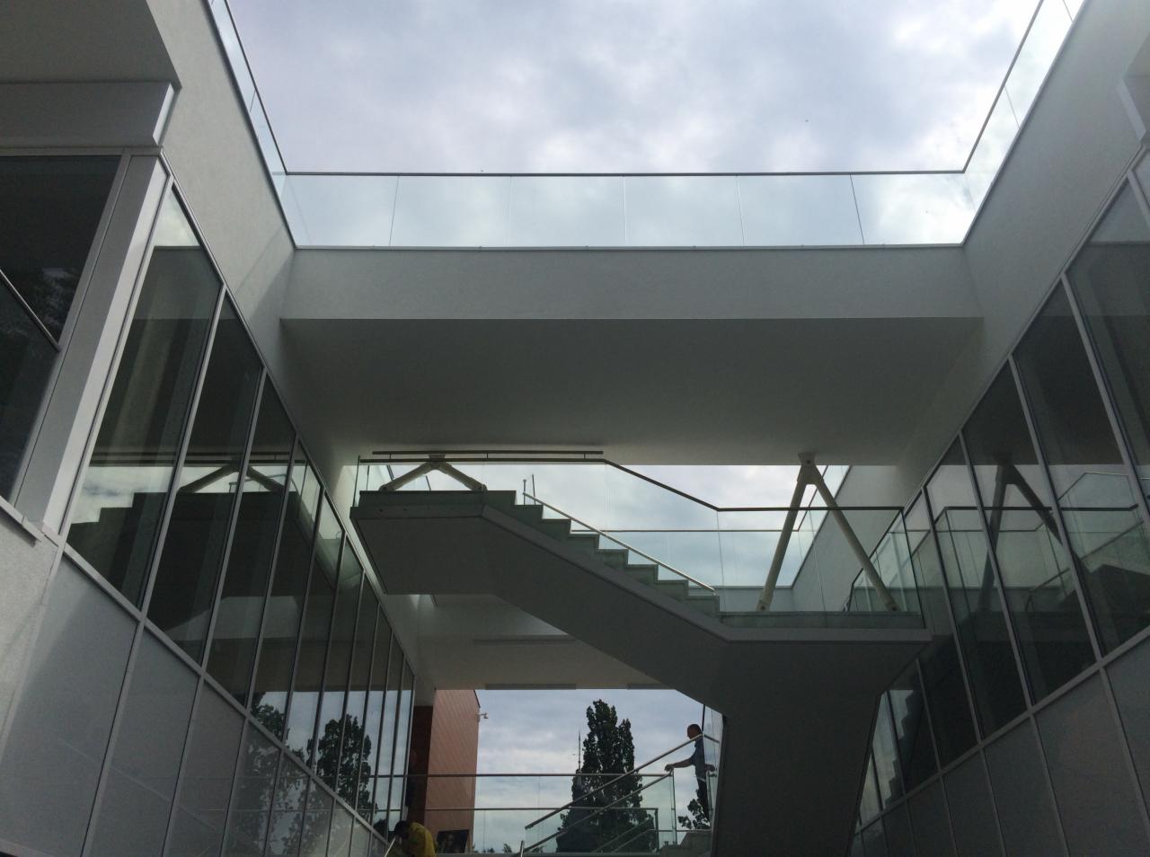 Nowoczesne schody z balustradami szklanymi wewnątrz budynku, widoczne z dołu, z elementami stalowymi i jasną kolorystyką, oświetlone naturalnym światłem z góry.