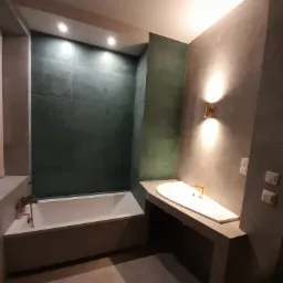 Nowoczesna łazienka z zielonymi płytkami w strefie kąpieli, minimalistyczną umywalką na betonowym blacie i złotą armaturą, oświetlona kinkietami.