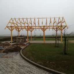 Konstrukcja drewnianej wiaty w trakcie montażu na zewnątrz, widoczne fundamenty pod słupy, kostka brukowa i mglista pogoda w tle.