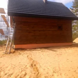 Brązowy drewniany dom z ciemnym dachem, jeszcze w trakcie budowy, ze rusztowaniem i drabiną z boku, widoczny fragment fundamentu z izolacją, teren wokół piaszczysty.