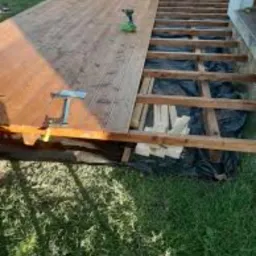 Montaż drewnianego tarasu: widoczne deski częściowo ułożone, konstrukcja nośna z czarną folią, wkrętarka akumulatorowa i zacisk stolarski na deskach.