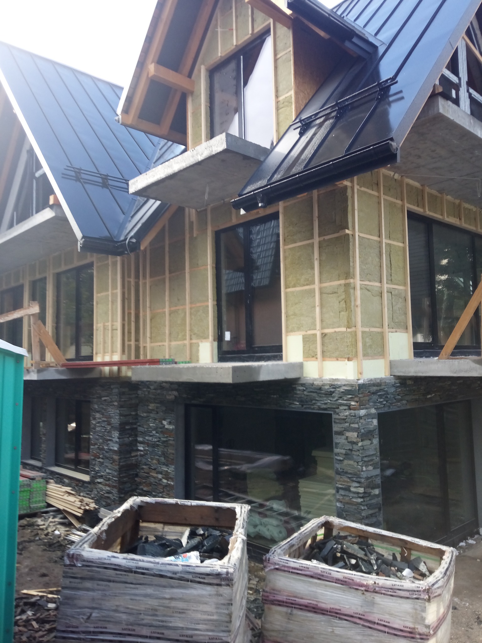 Budynek w trakcie budowy, z widoczną konstrukcją drewnianą, izolacją z wełny mineralnej i czarnym dachem z blachy na rąbek stojący, palety z materiałami budowlanymi w pierwszym planie.