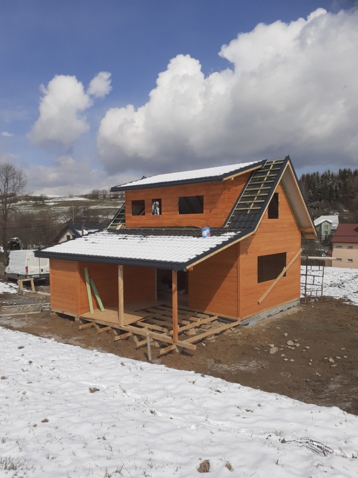 Surowy stan drewnianego domu z czarnym dachem w trakcie budowy, widoczne drewniane elementy konstrukcyjne tarasu i rusztowanie na dachu w zimowej scenerii.