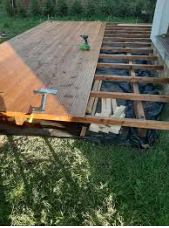 Montaż drewnianego tarasu: widoczne deski częściowo ułożone, konstrukcja nośna z czarną folią, wkrętarka akumulatorowa i zacisk stolarski na deskach.