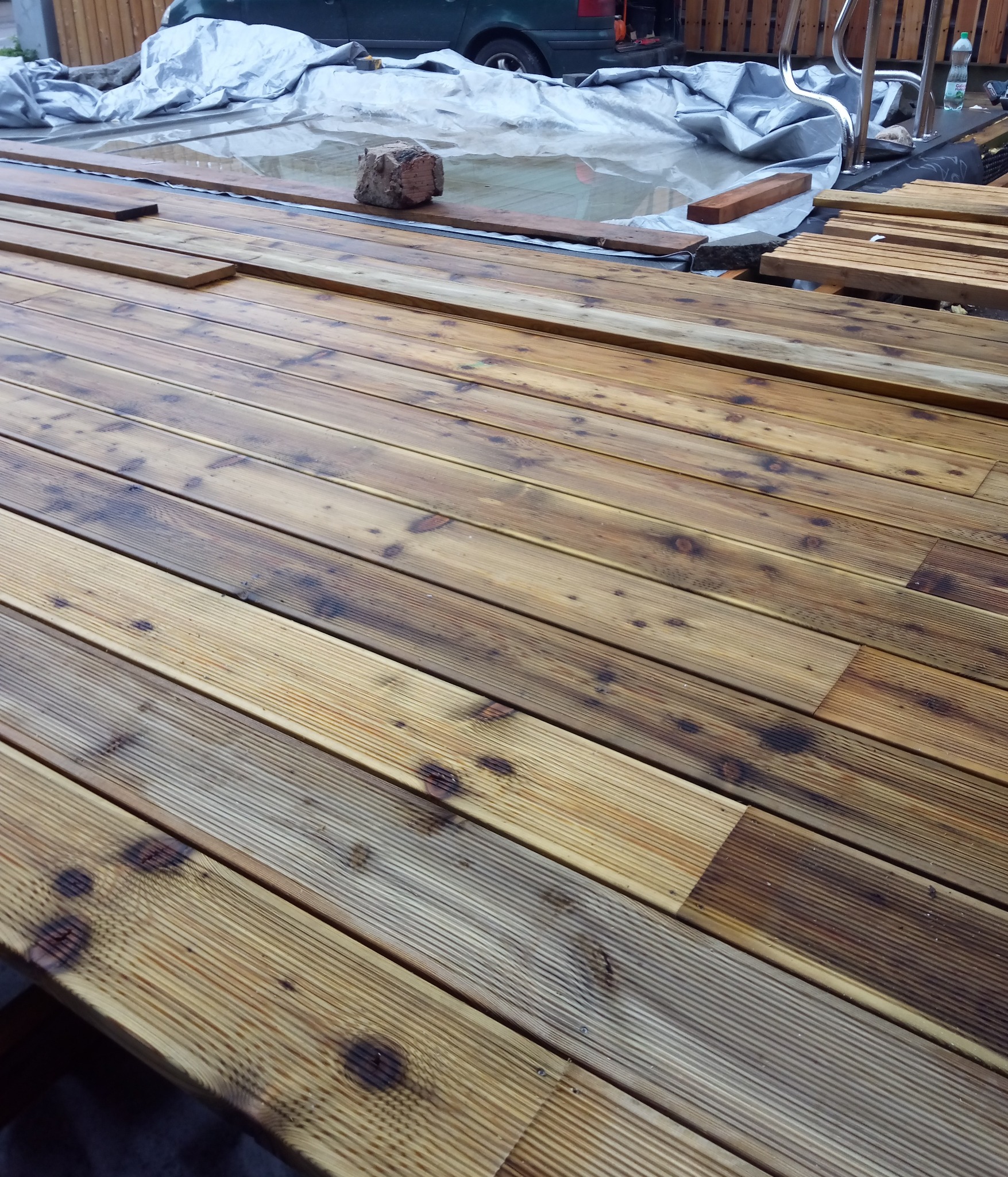 Montaż drewnianego tarasu przy basenie, deski częściowo ułożone, widoczny fragment basenu przykrytego plandeką i zaparkowany samochód w tle.