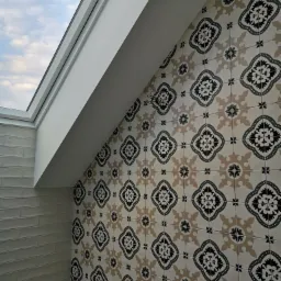 Ukośna ściana wyłożona dekoracyjnymi, wzorzystymi płytkami w odcieniach beżu i czerni, sąsiadująca z fragmentem ściany wyłożonej białymi, podłużnymi płytkami, oświetlona światłem wpadającym przez...